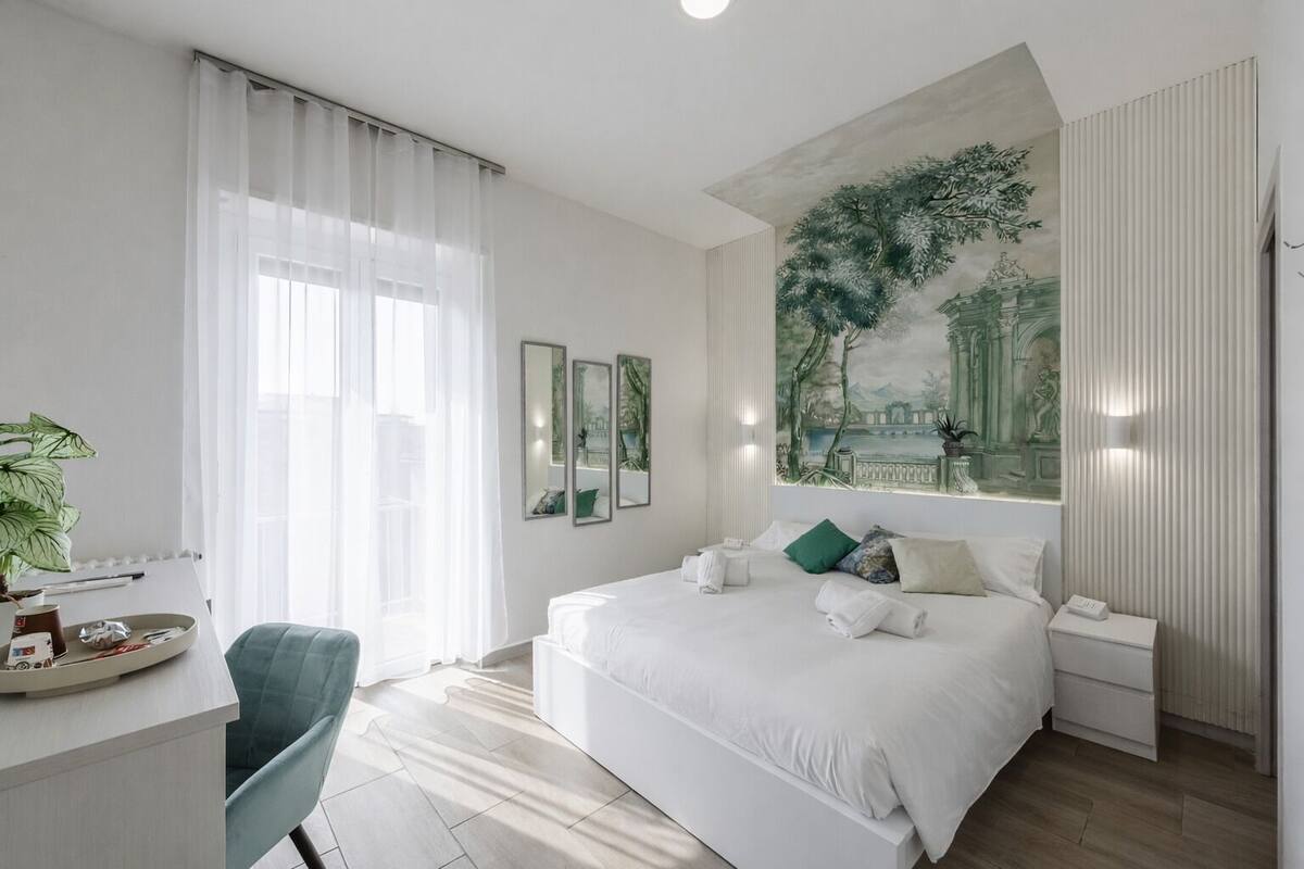 Een moderne hotelkamer met een groot bed, een bureau met stoel, een groot raam met dunne gordijnen, en een muurschildering.