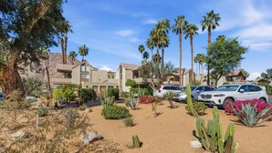 A desert-style residential area with multiple houses, palm trees, cacti, and a parking lot with cars. - Desert Escape With Private Balcony Condo (Indian Wells)