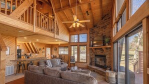 A spacious living area with a fireplace, a high ceiling with exposed beams, and a large window providing a view of the outdoors. - Grendel Getaway by Innsbrook Vacations! (Innsbrook)