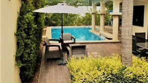 Un área de piscina con zona de estar techada, una palmera y un edificio con techo de tejas.