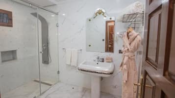 A bathroom with a glass-enclosed shower, a pedestal sink, a mirror with an ornate frame, and a robe rack with towels.
