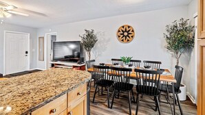 Una cocina moderna con encimeras de granito, un comedor con mesa de madera y sillas negras, un televisor de pantalla plana y dos plantas en macetas.