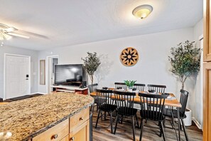 A modern kitchen with granite countertops, a dining area with a wooden table and black chairs, a flat-screen TV, and two potted plants. - 4BR Bio Hack Retreat: HTD Pool~Sauna~Hot Tub~Plunge (West Palm Beach)