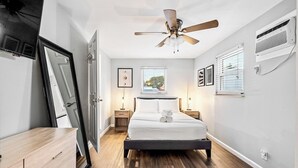 Un dormitorio con cama, ventilador de techo, ventana con persianas, mesita de noche y velador.