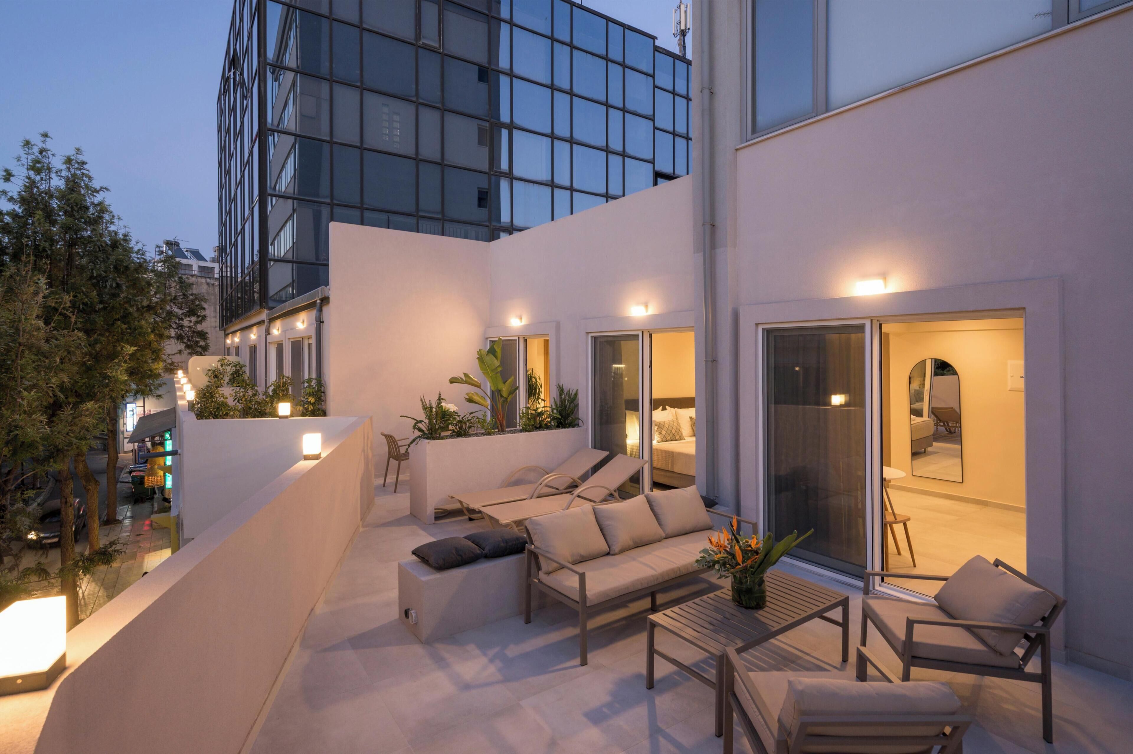 A modern outdoor living area with a sofa, chairs, and a table, surrounded by plants and a glass door leading to an interior space.