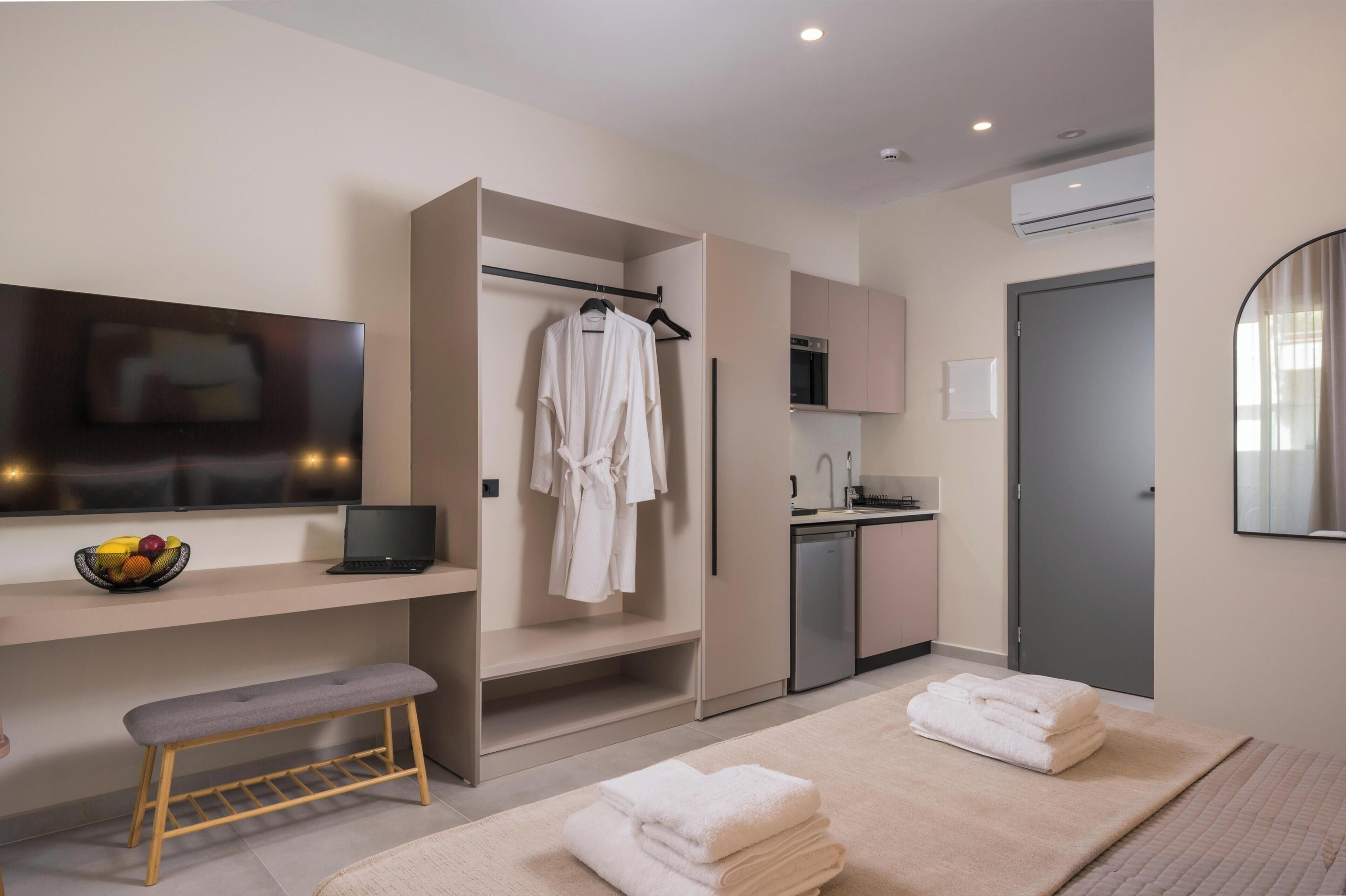 A modern hotel room with a TV, a laptop, a bench, a robe, a sink, and a door.