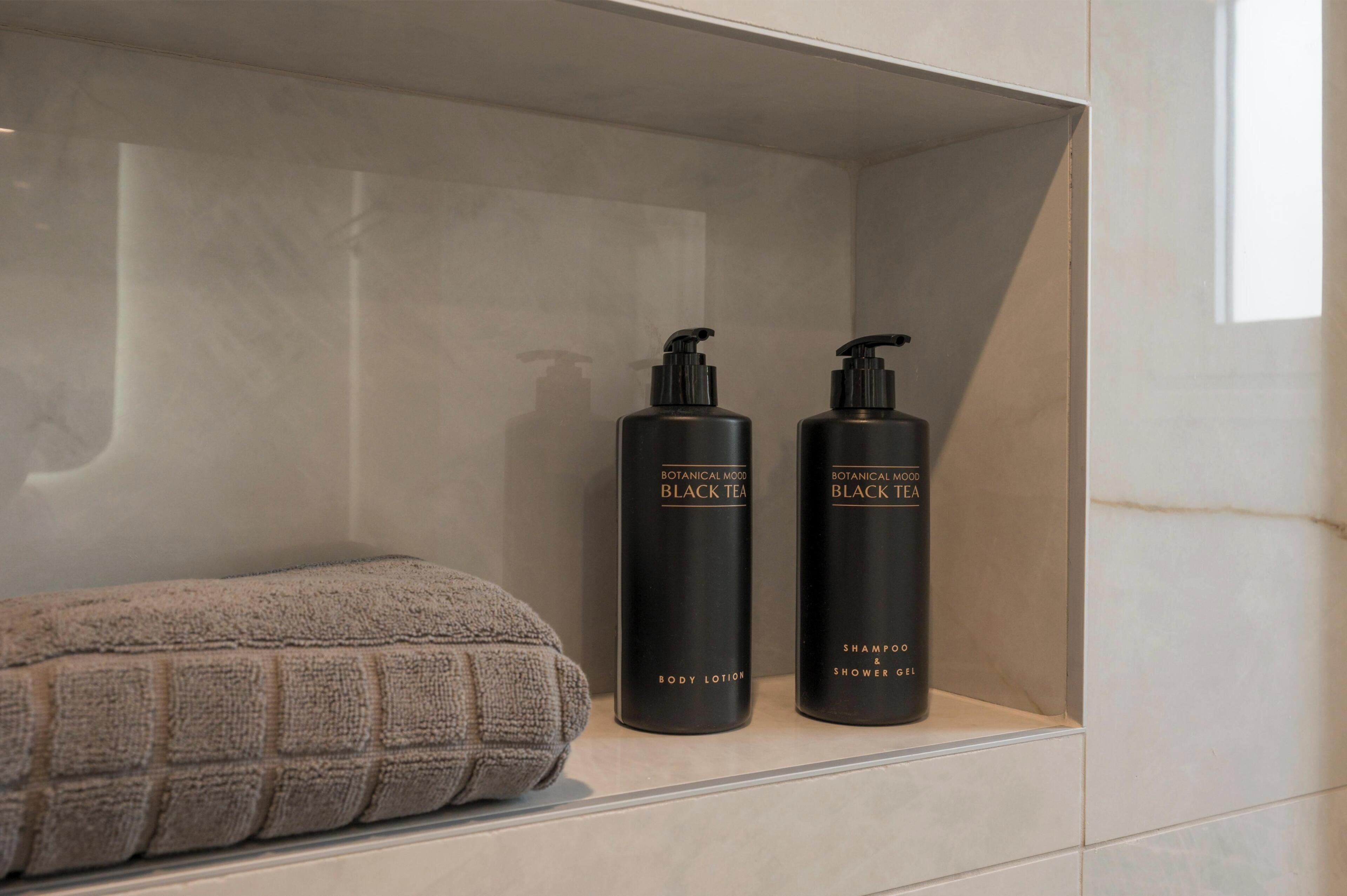 Uno scaffale in bagno con due flaconi neri etichettati 'Botanical Mood Black Tea Body Lotion' e 'Shampoo & Shower Gel', e un asciugamano a trama.