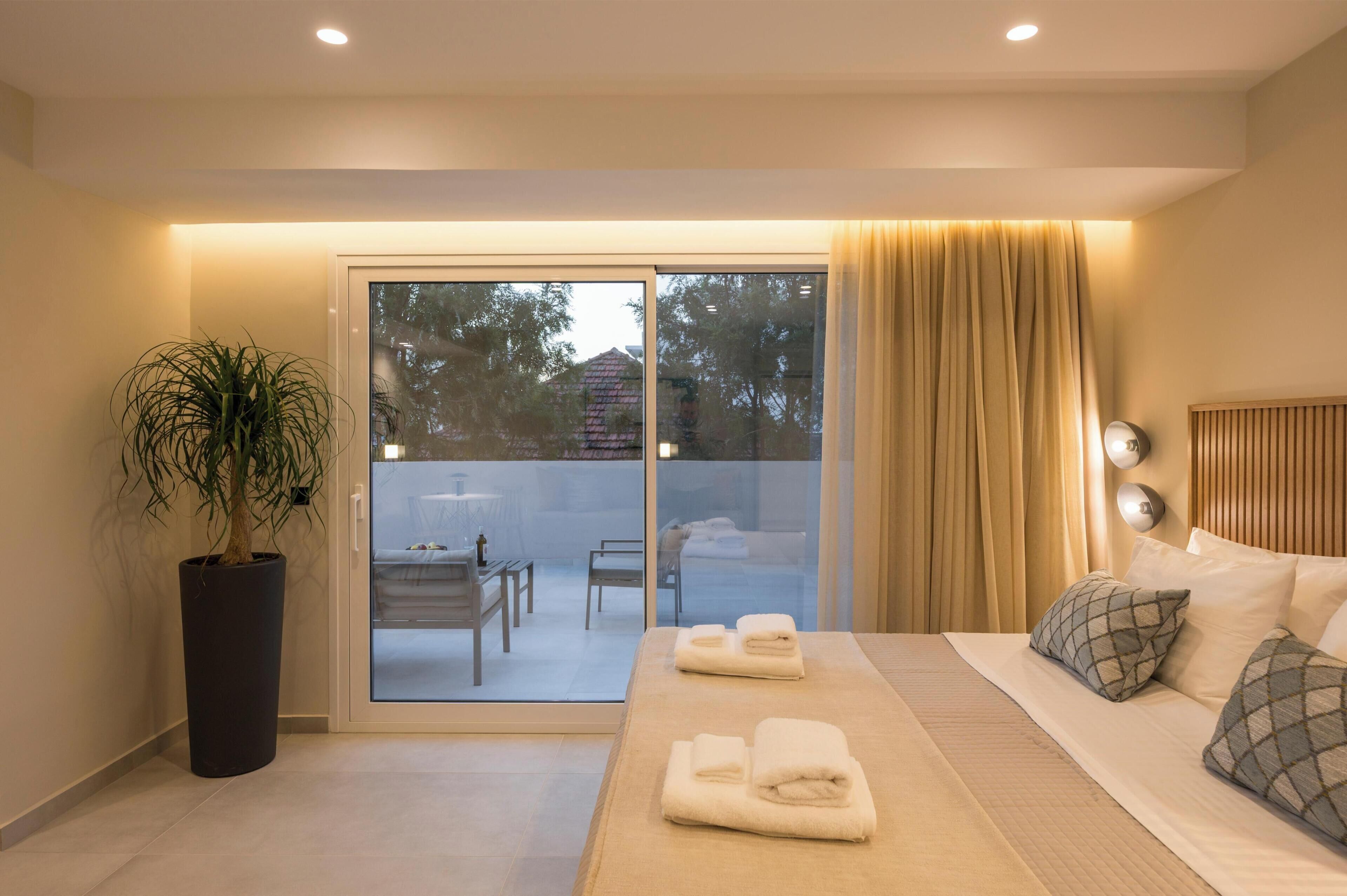 A modern hotel room with a bed, a potted plant, a sliding glass door to a balcony, and a view of the outside.