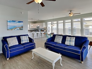 A living room with two blue sofas, a white coffee table, and a kitchen in the background. - Searenity On The Sound | Sound Views, Private Dock + Luxury Amenities (Surf City)