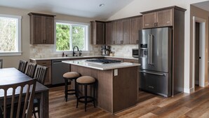 A modern kitchen with wooden cabinets, a large island, stainless steel appliances, and a dining area with bar stools. - May Creek Peaks: Custom built home on private acres w/ mountain views, covered spa, luxury amenities (Gold Bar)