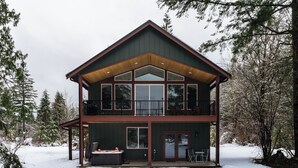 A two-story cabin with a balcony, large windows, and a hot tub. - May Creek Peaks: Custom built home on private acres w/ mountain views, covered spa, luxury amenities (Gold Bar)