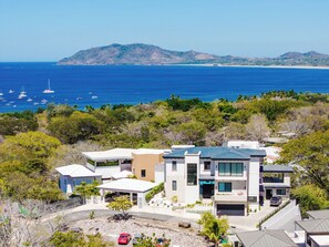 A modern multi-story house with a garage, surrounded by lush greenery and a view of the ocean.