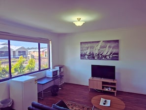 Een woonkamer met een groot raam, een flatscreen televisie, een schilderij van zeilboten, een houten salontafel en een klein bureau met een computer.