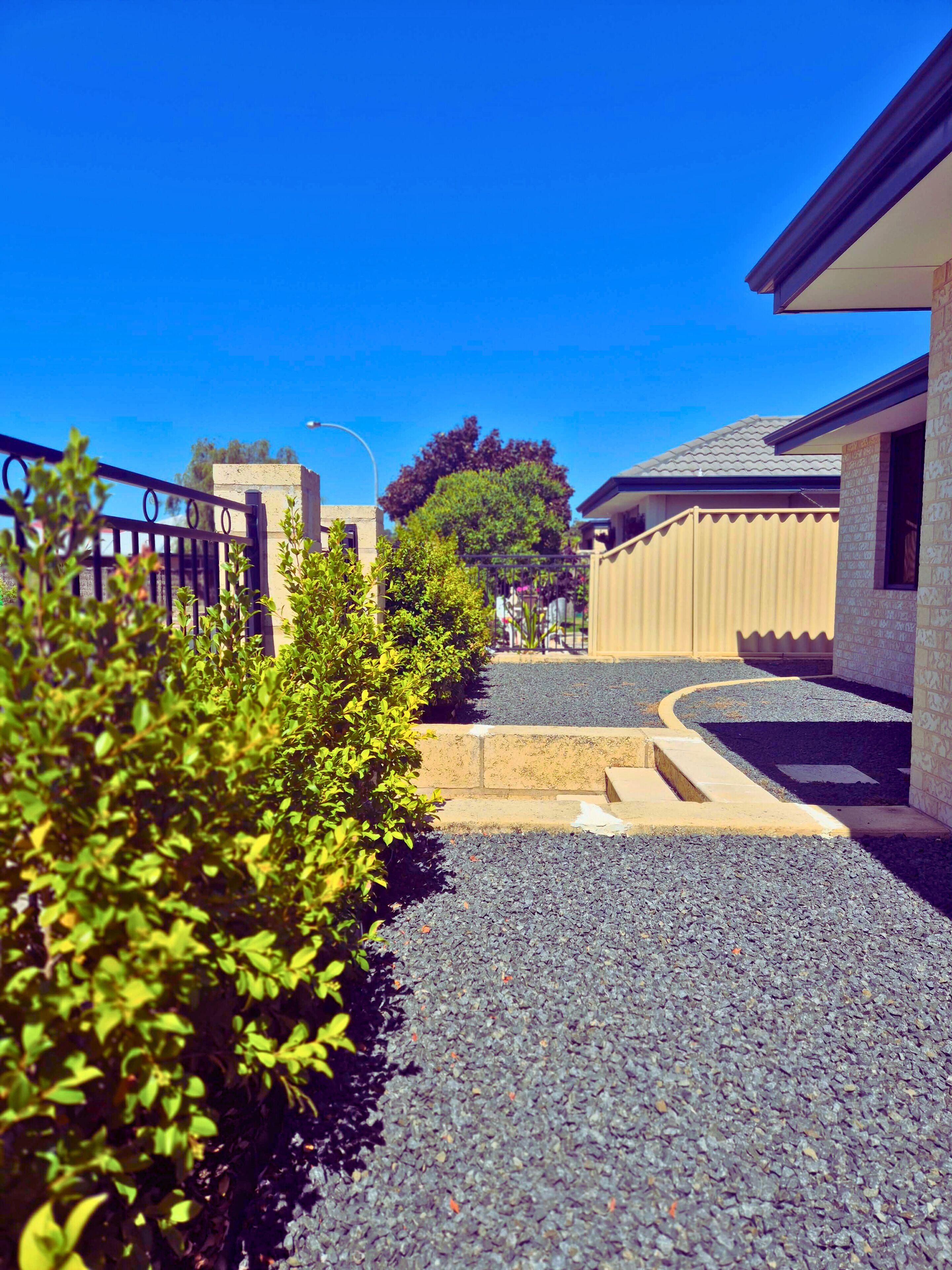 A residential street with a gravel driveway, a metal fence, and houses with brick and stucco exteriors.