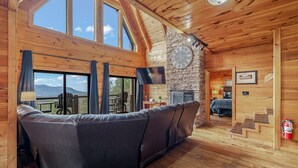 Un interior de madera con sofá, chimenea de piedra y un ventanal con vistas panorámicas.
