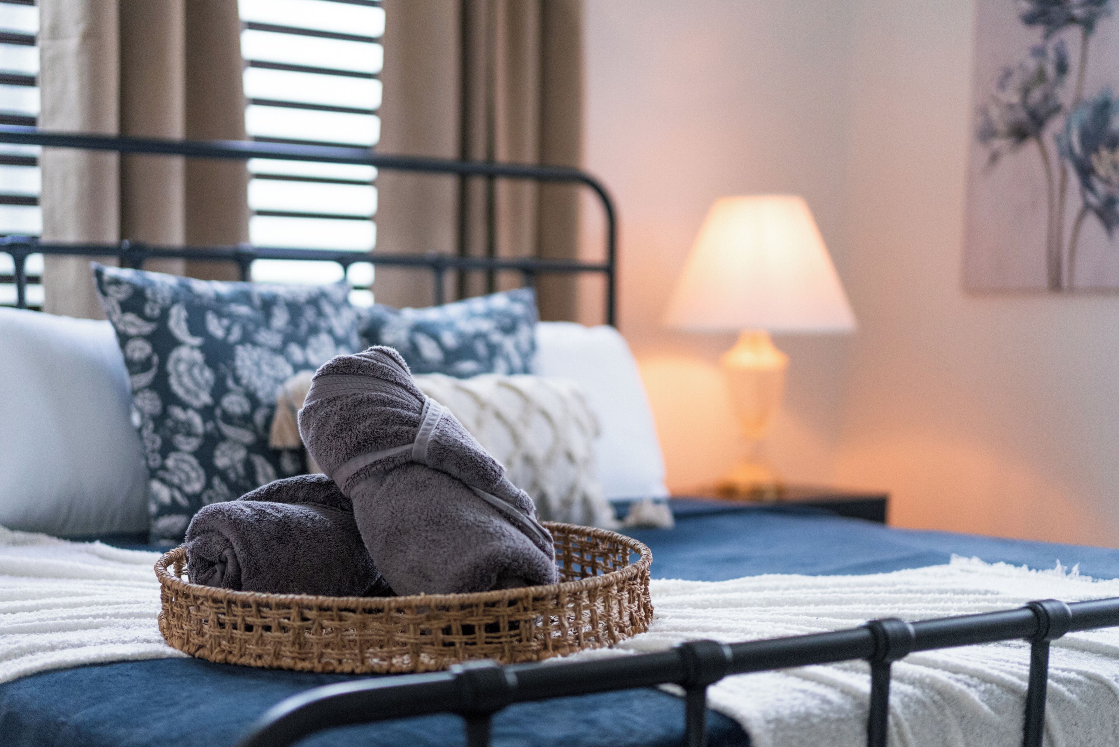 A neatly made bed with a basket of folded towels, a bedside lamp, and a floral painting on the wall.