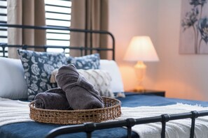 Una cama bien hecha con una cesta de toallas dobladas, una lámpara de noche y un cuadro floral en la pared.
