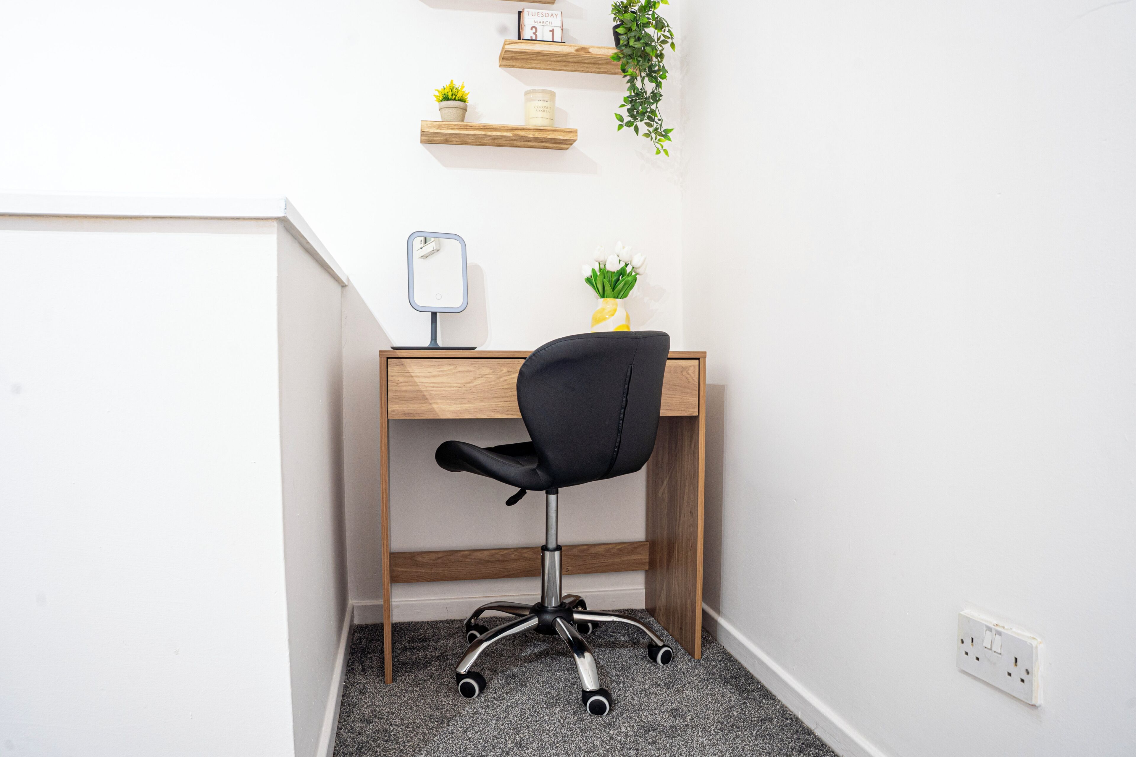 Ein Büro im Eck mit Schreibtisch aus Holz, schwarzem Bürostuhl, kleinem Spiegel, Topfpflanze und Wanduhr.
