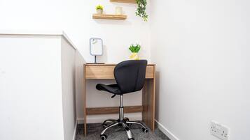 Ein Büro im Eck mit Schreibtisch aus Holz, schwarzem Bürostuhl, kleinem Spiegel, Topfpflanze und Wanduhr.