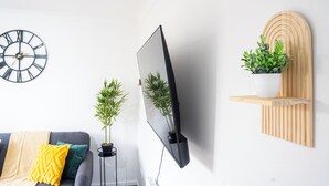 Ein modernes Wohnzimmer mit einem wandmontierten Flachbildfernseher, einer runden Wanduhr, einem grauen Sofa mit Kissen und zwei Blumentöpfen.