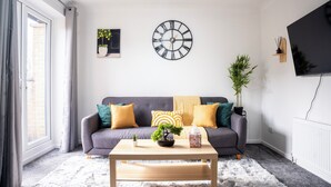 Ein Wohnzimmer mit einer Couch, einem Couchtisch, einer Uhr, einem Fernseher und einem Fenster mit Vorhängen.