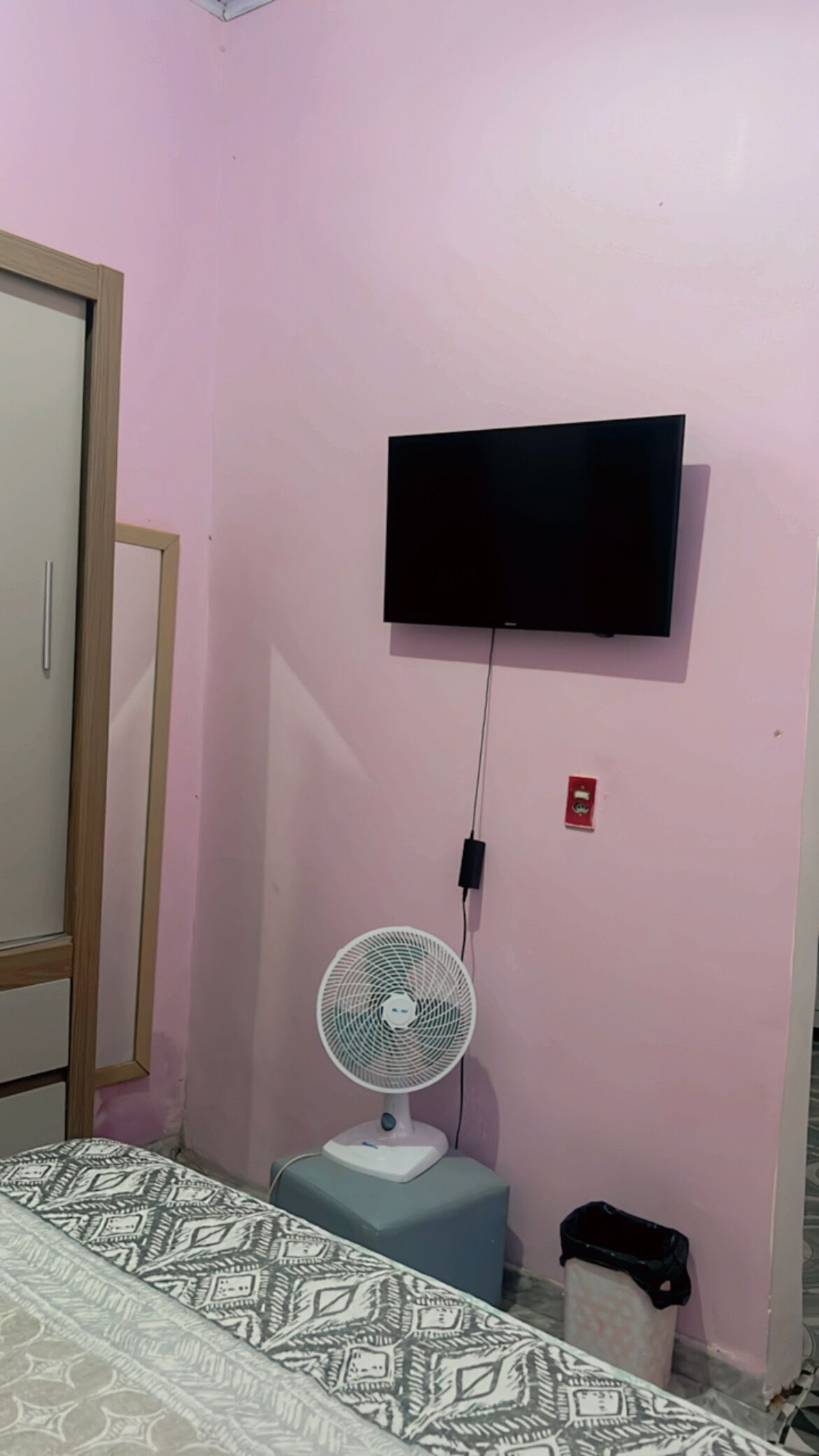 Una habitación con una cama, un televisor de pared, un ventilador de pie y un cubo de basura.
