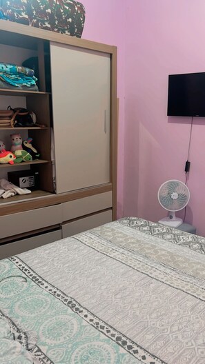 Ein Schlafzimmer mit Kleiderschrank, einem Bett mit gemusterten Betttextilien, einem an der Wand montierten Fernseher und einem Standventilator.