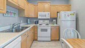 A compact kitchen with wooden cabinets, a microwave, a stove, a refrigerator, and a dining area with a chair. - 3 Bedroom House (7033) in Ocean Lakes Campground (Myrtle Beach)