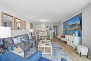 A modern living room with a sofa, coffee table, and a dining area with chairs. - Ocean Creek North H7 - Pools (Myrtle Beach)