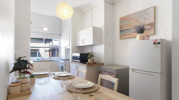 A modern kitchen with white cabinets, a dining table set with plates and cutlery, a hanging light fixture, and a painting on the wall.