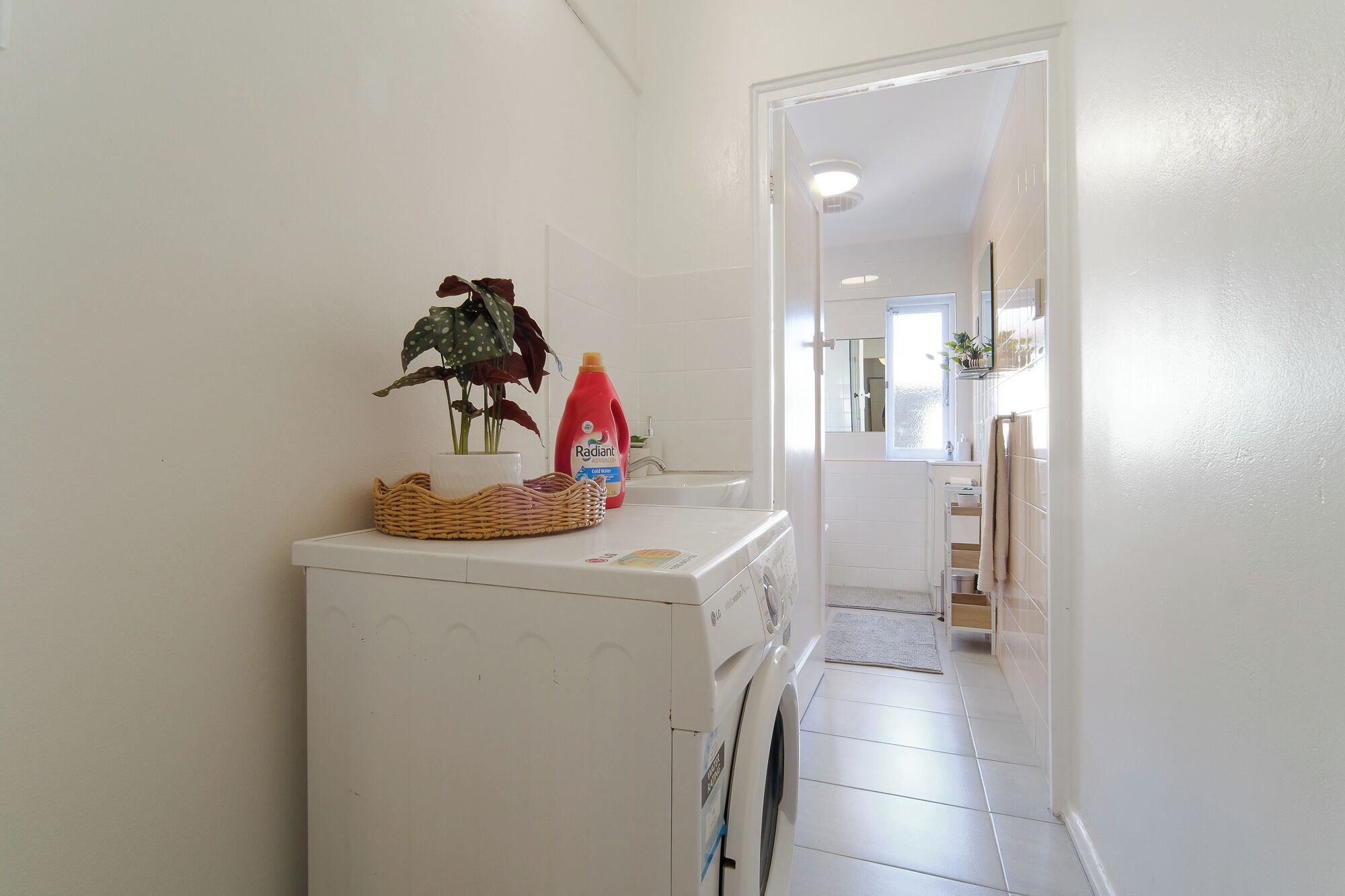 A laundry area with a washing machine, a basket, a bottle of detergent, and a potted plant.