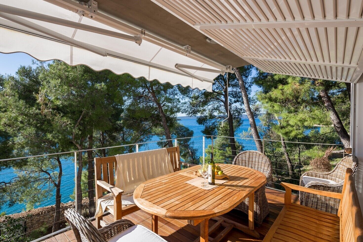 Una terrassa de fusta amb una taula rodona i cadires de vimet, amb vista a una zona costanera amb arbres i el mar.