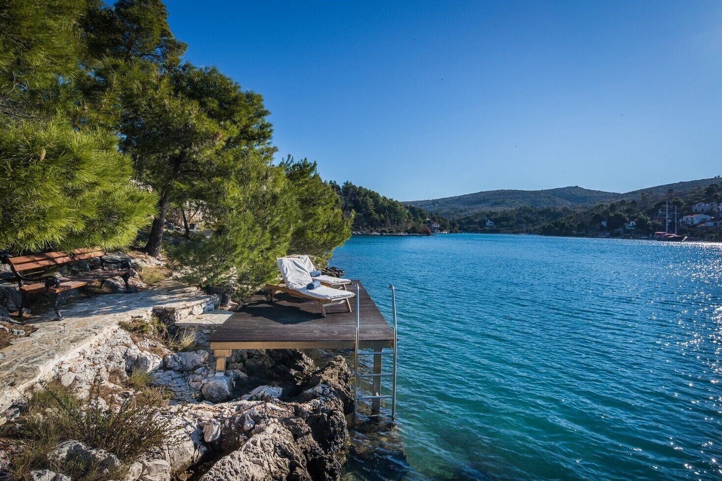 A wooden deck with a bench and lounge chairs overlooking a clear blue lake, surrounded by trees and rocky terrain.