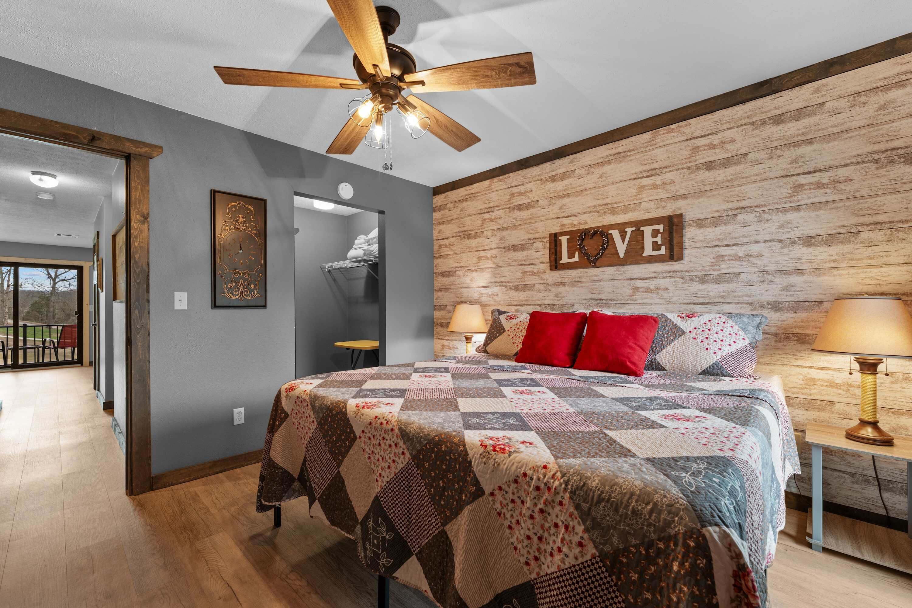A bedroom with a quilted bedspread, a ceiling fan, a wooden wall with a 'LOVE' sign, and a view into the adjacent room.