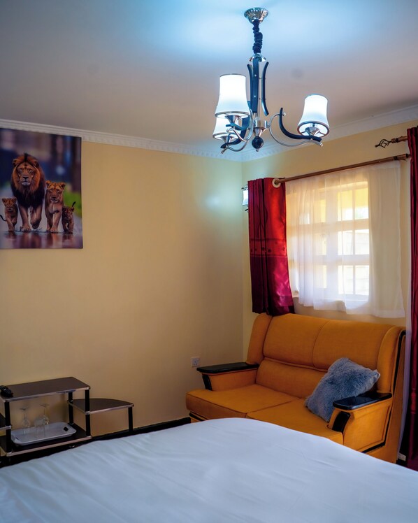 Une chambre d’hôtel avec un lit, un canapé, un bureau, une lampe, un tableau représentant des lions et une fenêtre avec des rideaux.