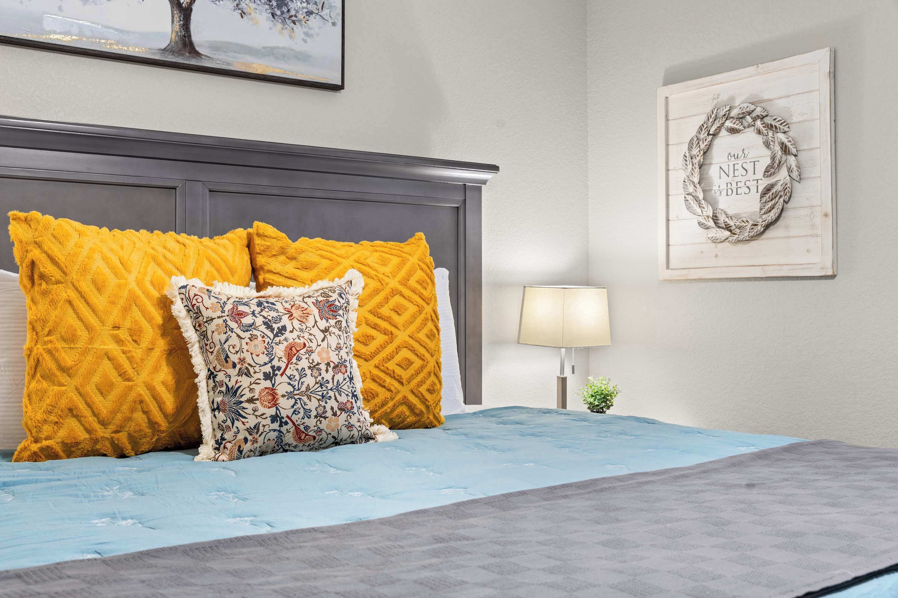 Una cama con una colcha azul, dos almohadas mostaza y una almohada floral.