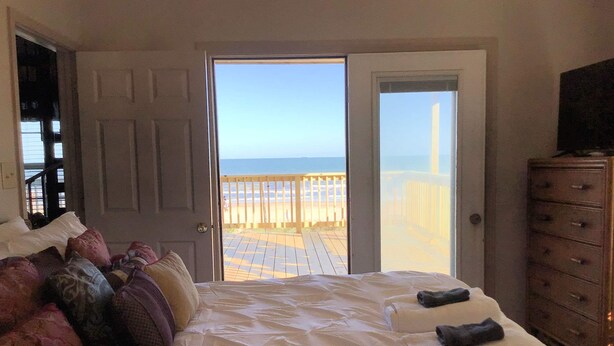 Een slaapkamer met een bed, kussens, een dressoir en een deur die uitkomt op een balkon met uitzicht op het strand.