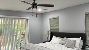 Ein Schlafzimmer mit Bett, Deckenventilator, Nachttisch, Lampe und Fenster mit Jalousien.