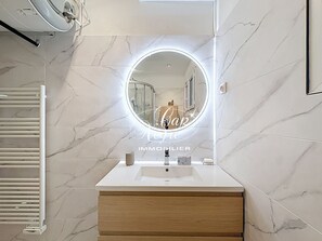 A bathroom with a marble wall, a round mirror with a light, a white sink, and a wooden cabinet. - Elegant family villa with sea view for 4 people at La Fossette (Le Lavandou)