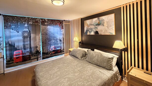 Une chambre à coucher avec un lit, un tableau abstrait accroché au mur, une lampe et une fenêtre donnant sur un paysage urbain.