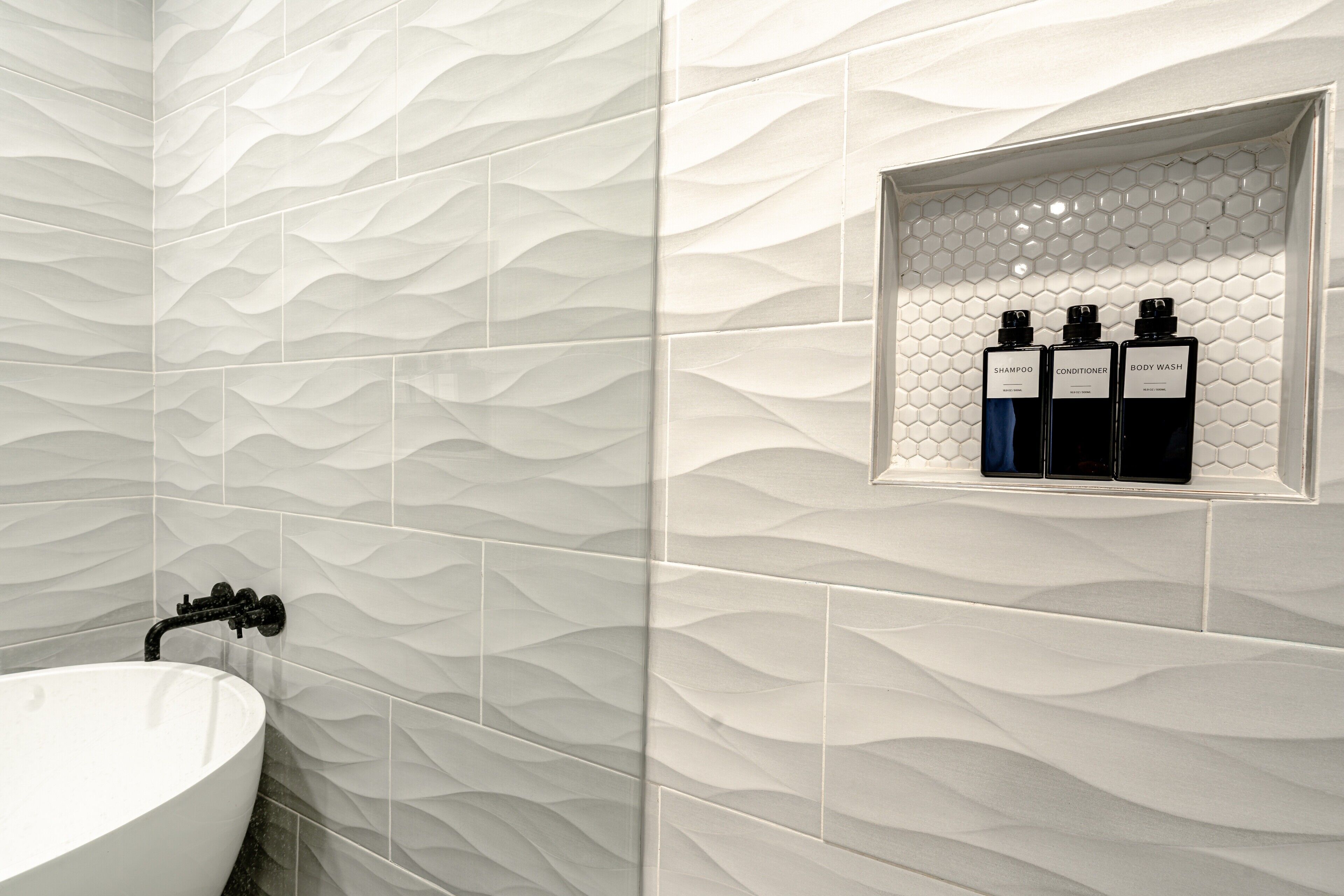 Un bagno con una parete bianca a texture, un lavandino bianco e tre flaconi etichettati shampoo, balsamo e bagnoschiuma.