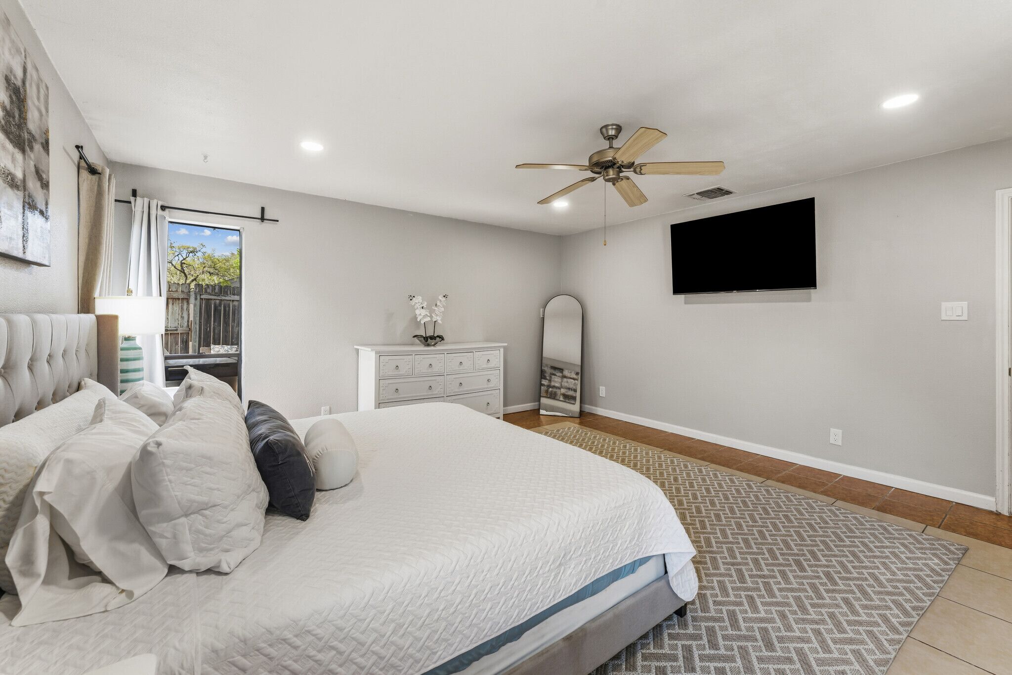 Une chambre à coucher comprenant un lit, un ventilateur de plafond, une télévision, une commode, un miroir et une fenêtre avec des rideaux.