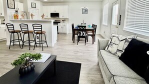A modern kitchen with white cabinets, a dining area with a table and chairs, and a living room with a sofa and a coffee table. - Big Beautiful BOLD (Grand Prairie)