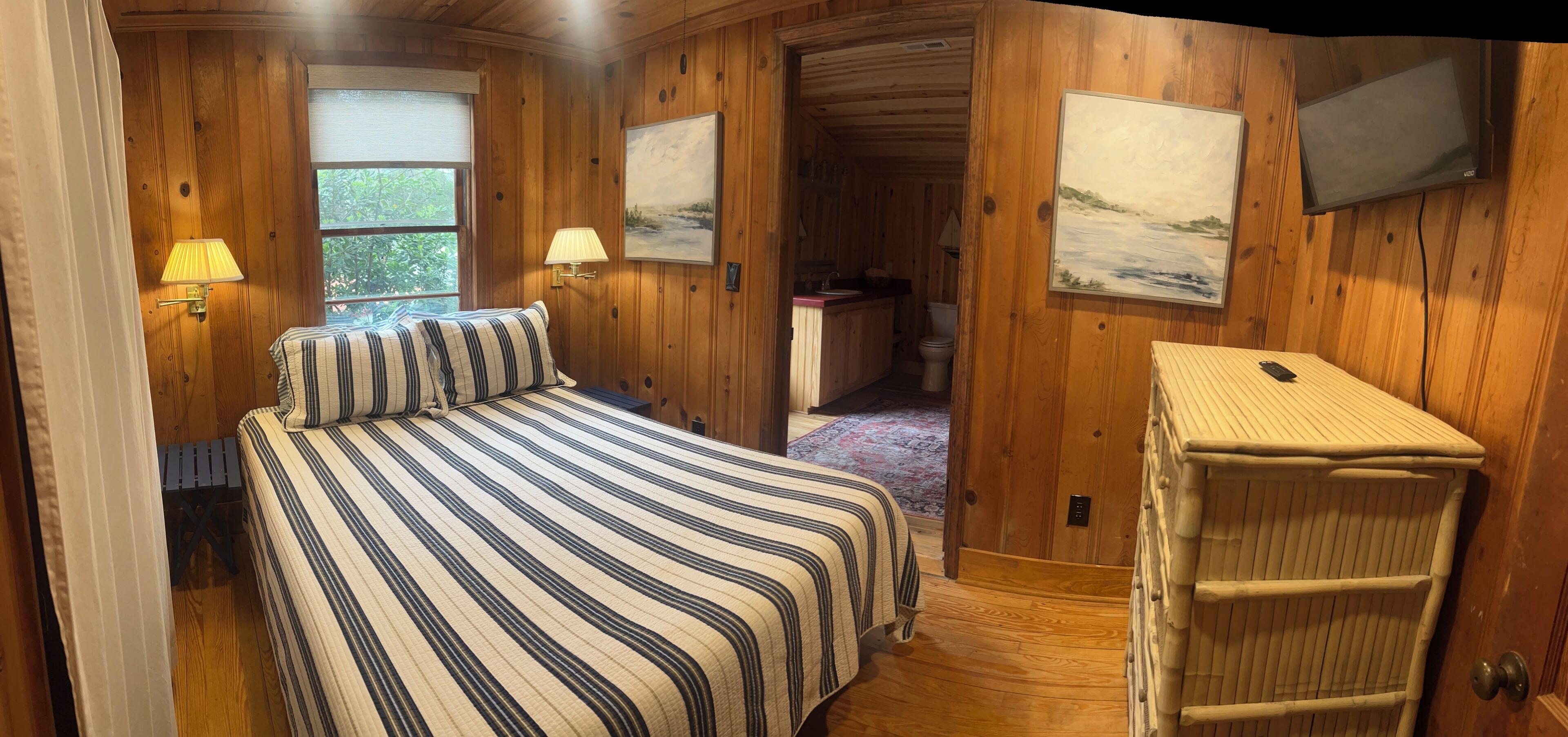 A wooden cabin bedroom with a bed, two wall-mounted lamps, a television, and a small bathroom visible through an open door.