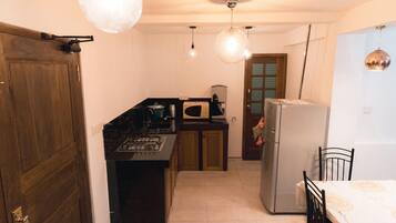 A compact kitchen with a stove, microwave, and refrigerator. There is a wooden door on the left and a dining area with a table and chairs on the right.