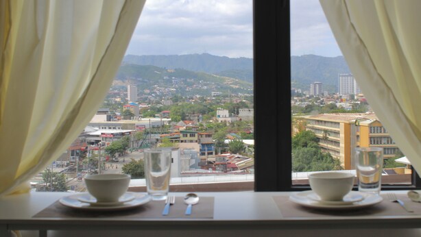 A table with a bowl, spoon, and glass set against a window with curtains, overlooking a cityscape. - Pure Simplicity: Sip Coffee Overlooking the Glittering City (Cebu City)