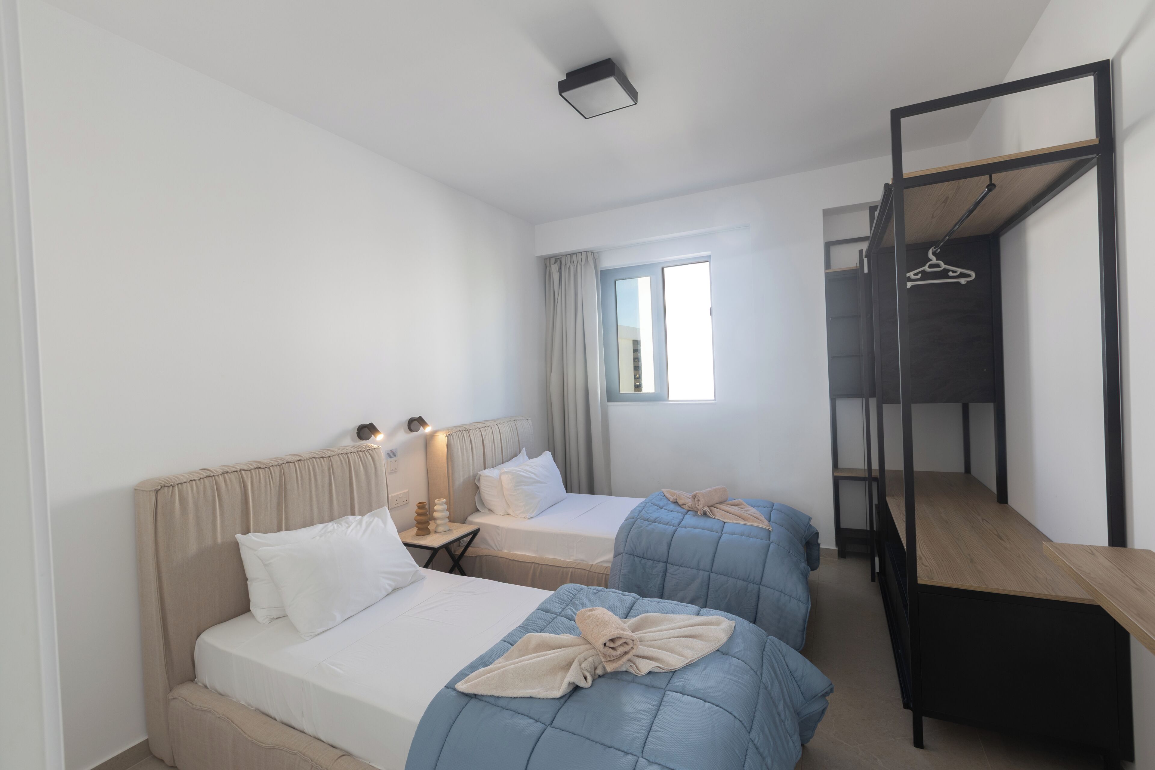 A room with two beds, a desk, a hanging rack, and a window with curtains.