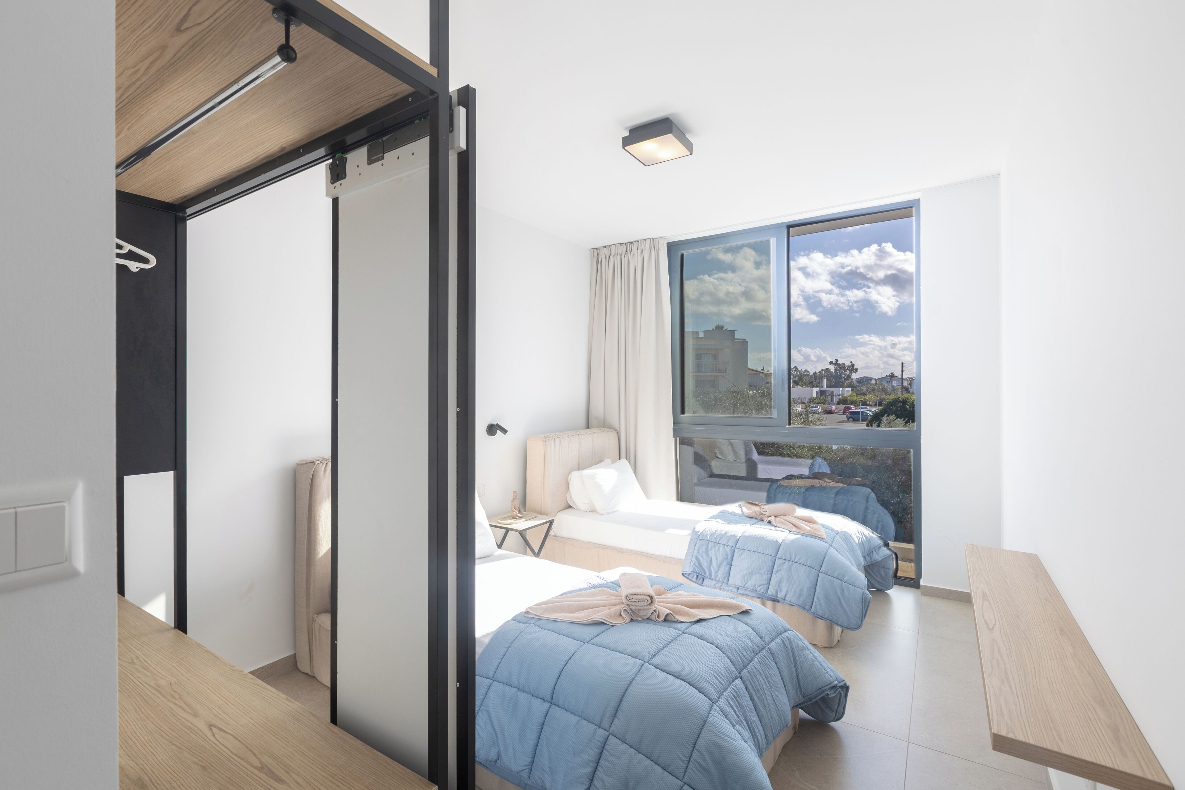 Una habitación de hotel moderna con puerta corredera, un ventanal grande con vista a la ciudad, dos camas con ropa de cama azul, una mesita de noche y un pequeño escritorio con silla.