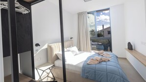 A modern bedroom with a bed, a small table, a window with curtains, and a view of the outside. - Ayia Napa Orea Villa OR6 (Ayia Napa)