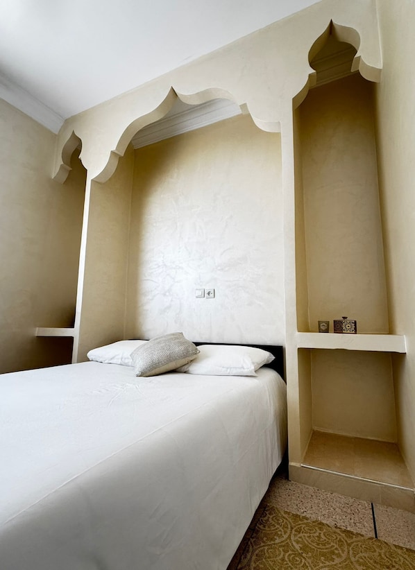 A bedroom with a bed, pillows, a nightstand, a shelf with decorative items, and a patterned rug.